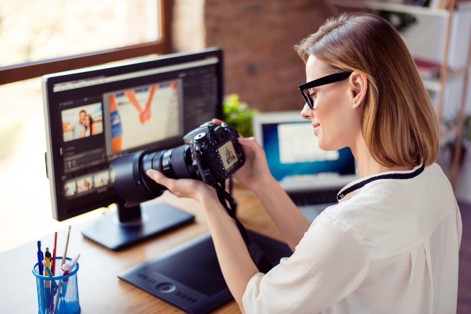 Side view of female freelancer photographer checking photos on a digital camera while sitting at the table in workstation